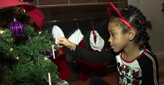 A young girl wearing a red headband decorates a Christmas tree with a candy cane; stockings are hanging on a fireplace in the background.