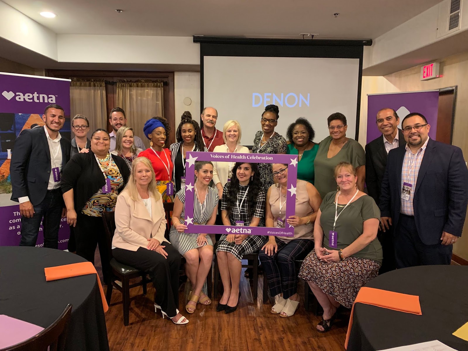 A diverse group of people poses together indoors at an Aetna-sponsored event, with some holding a purple frame labeled “Voices of Health Celebration.”.