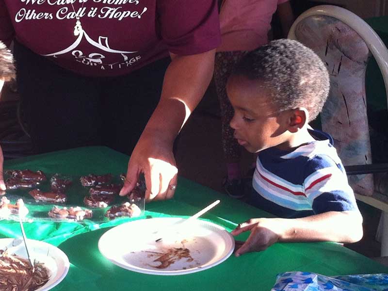 A child in a striped shirt sits at a table with a paper plate, while an adult arranges treats on the table.