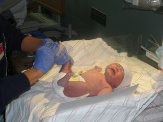 A newborn baby lies on a hospital blanket while a person wearing blue gloves tends to them in a medical setting.