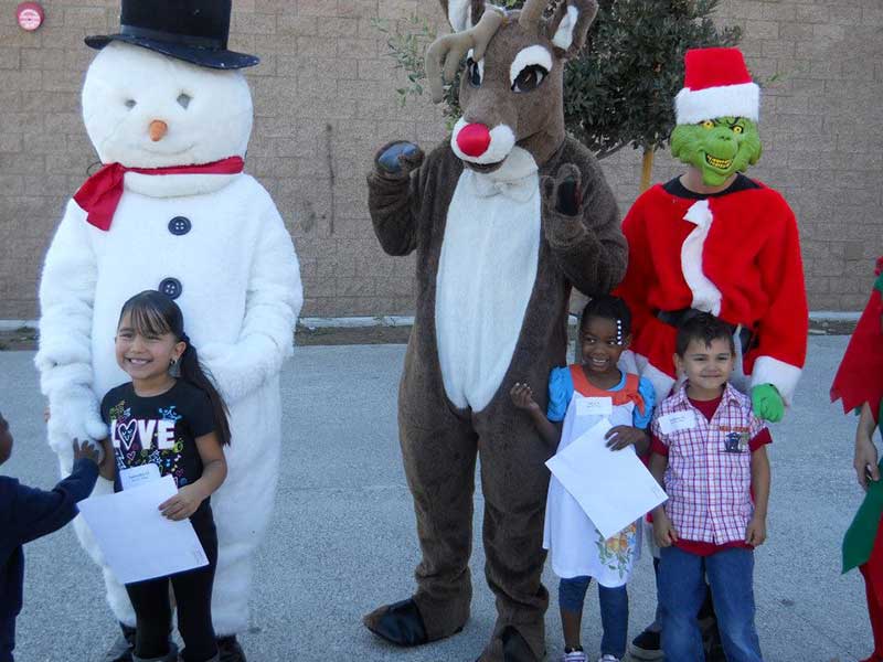 Three children stand in front of people dressed as a snowman, reindeer, and the Grinch. The children are holding papers and smiling outdoors.