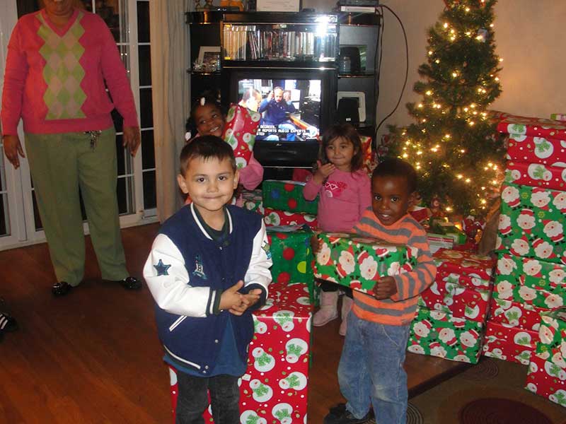 Four young children holding wrapped presents stand near a small decorated Christmas tree, with a woman and a TV in the background.