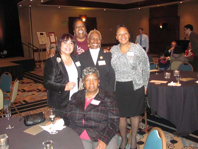 Five women pose together and smile at a round table in a banquet hall, with other people and tables visible in the background.