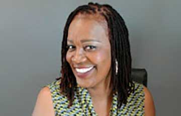 Smiling woman with medium-length, dark dreadlocks, wearing a patterned sleeveless top, sitting in front of a plain gray background.