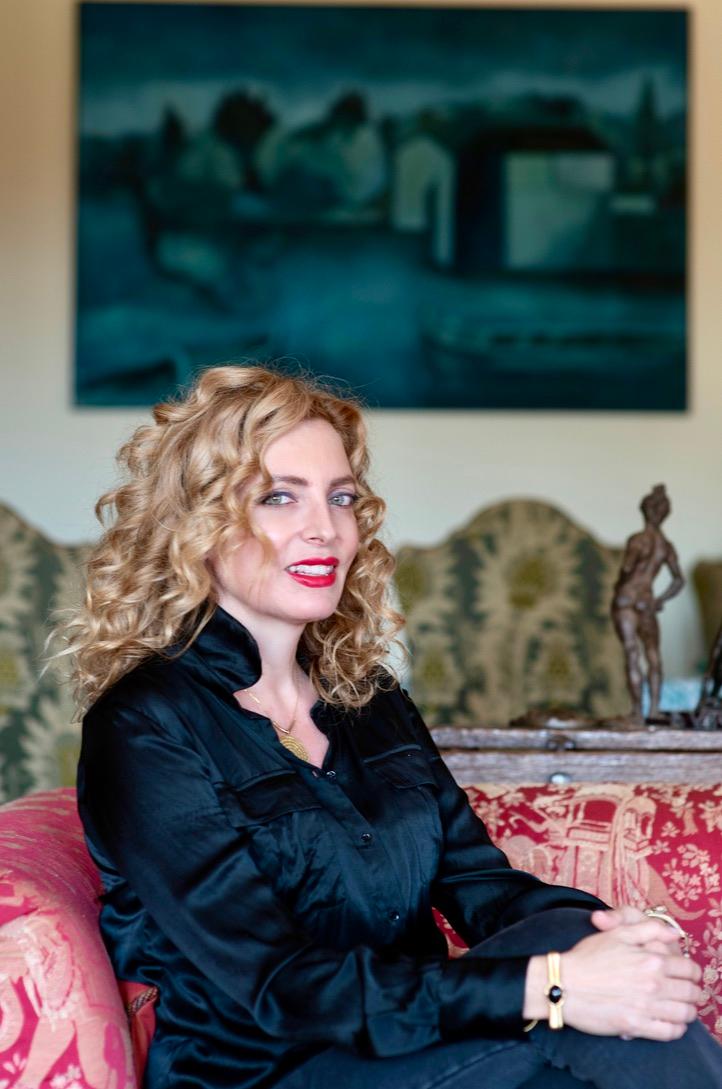 A woman with curly blonde hair and red lipstick sits on a patterned sofa, wearing a black blouse. There is a painting and a small sculpture in the background.