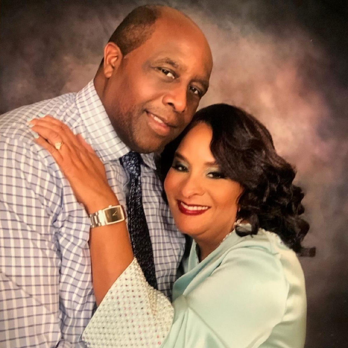 A man in a checkered shirt and tie is embraced by a smiling woman in a light blue top, both posing closely in front of a mottled studio background.