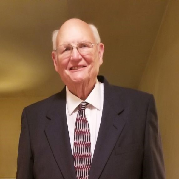 An older man with glasses, dressed in a dark suit, white shirt, and patterned tie, smiles while standing indoors against a plain beige wall.