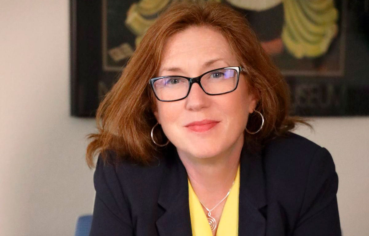 A woman with red hair and glasses, wearing a dark blazer and yellow top, sits indoors and looks at the camera.