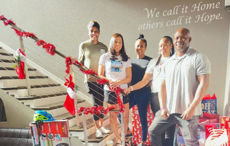 Five people stand together on a decorated staircase with Christmas stockings and gifts, under a wall sign that reads, "We call it Home, others call it Hope.