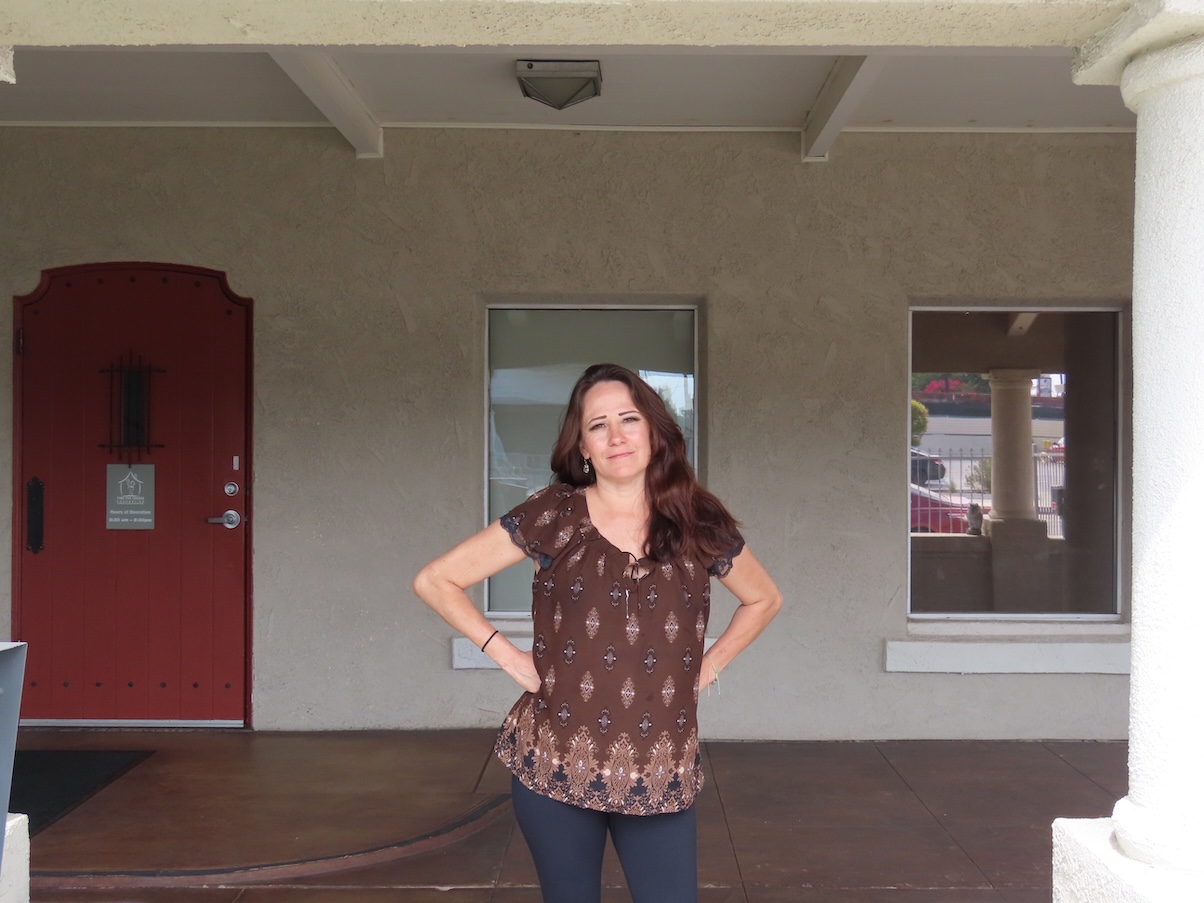 A woman with long brown hair stands with hands on hips in front of a beige building with a red door and two windows.