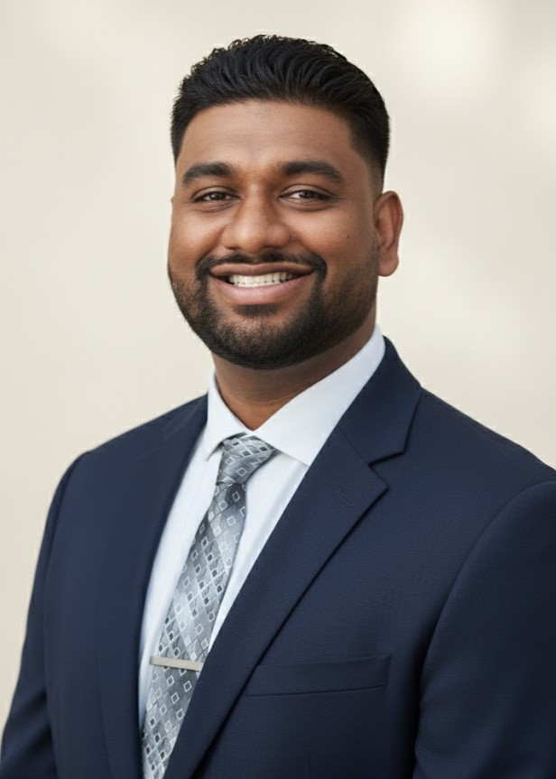 A man in a navy blue suit, white shirt, and patterned tie smiles at the camera against a neutral background.