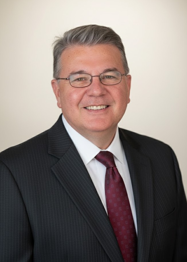 Smiling man wearing glasses, a dark pinstripe suit, white shirt, and red tie, posing for a professional headshot against a plain light background.