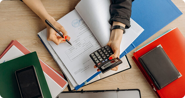 Person using a calculator and writing on documents at a desk with folders, a notebook, and a smartphone.