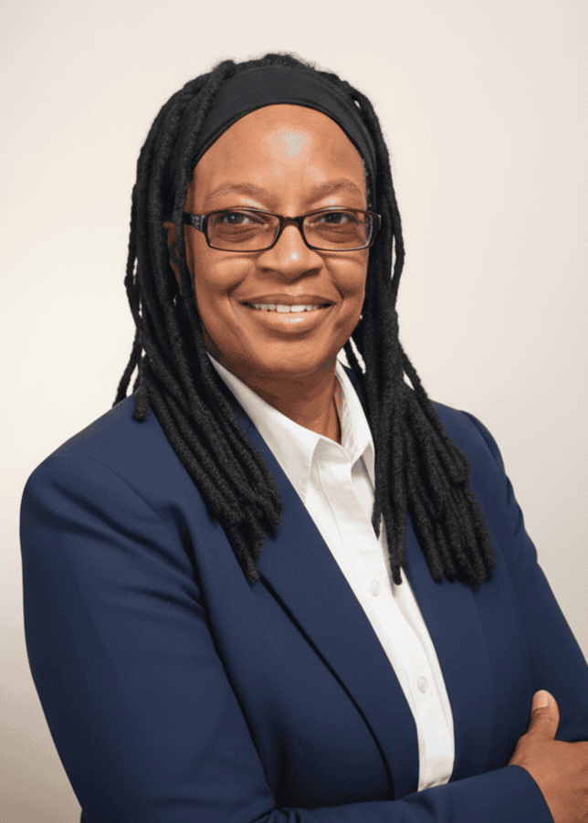 A person wearing glasses, a navy blazer, and a white shirt is smiling with arms crossed against a plain light background.