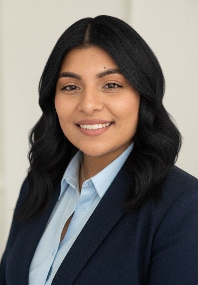 Woman with long dark hair wearing a navy blazer and light blue shirt, smiling at the camera against a plain, light background.