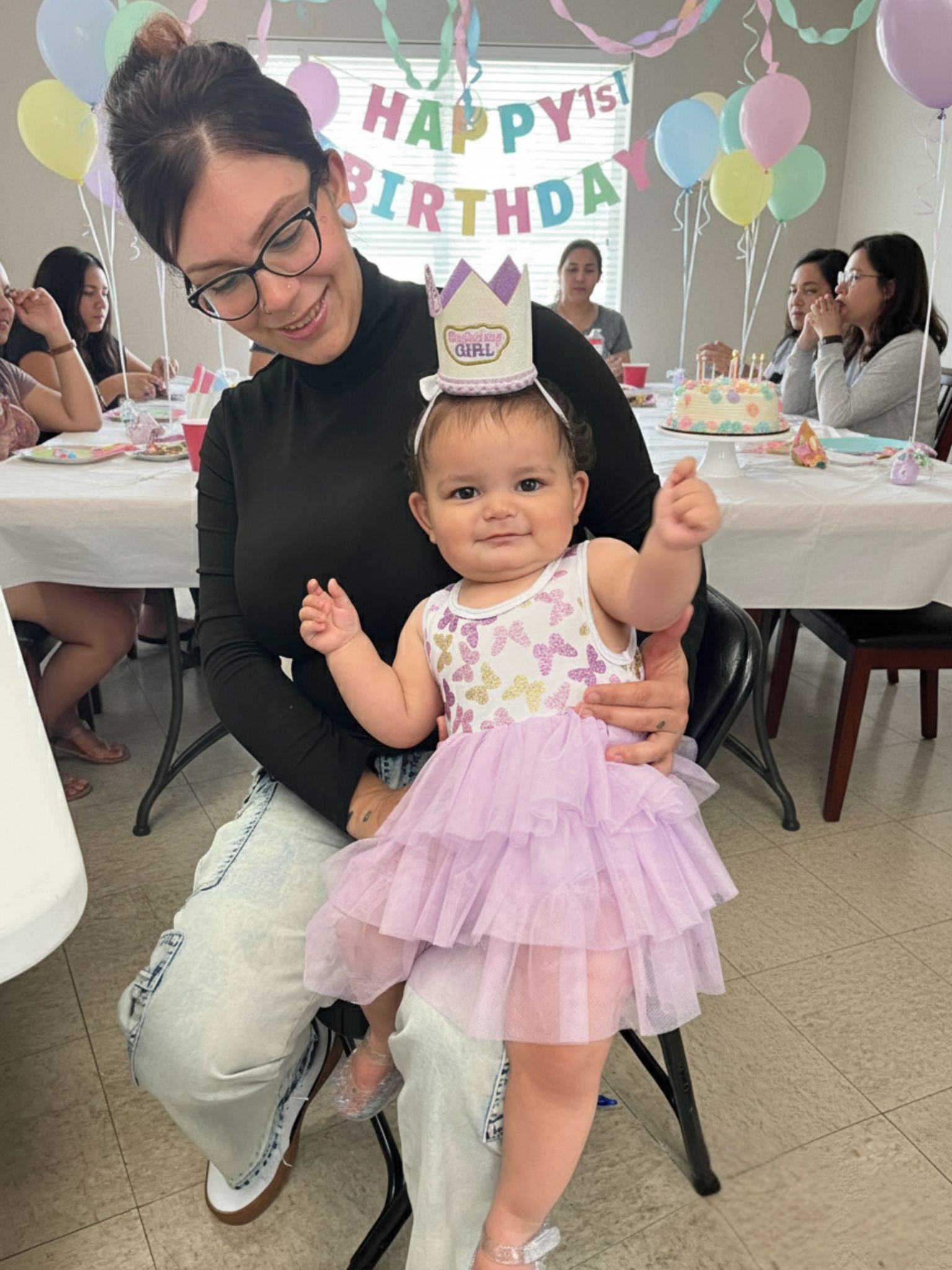 A woman sits holding a smiling baby wearing a "Birthday Girl" crown at a decorated birthday party with balloons and a "Happy 1st Birthday" banner.